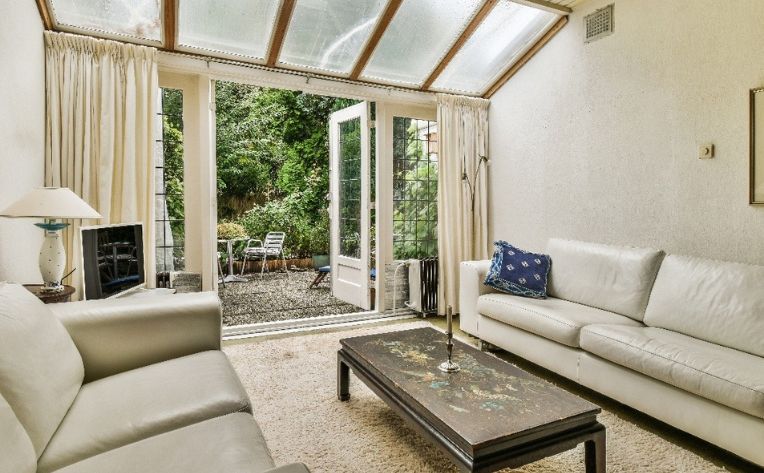 Alt TextBright sunroom living area with white leather sofas, glass ceiling, coffee table, and open French doors leading to a garden patio.